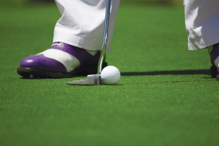 Close-up of a golfer's shoes and a golf ball near a putter on a green golf course, highlighting golf equipment and techniques.