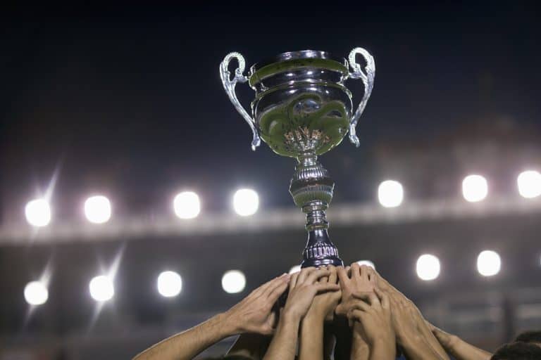 Shiny silver trophy being lifted by multiple hands at a nighttime sports event, celebrating victory and achievement, illuminated stadium lights in the background.