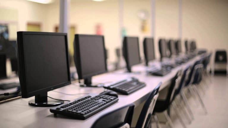 Desktop computers and keyboards on a long row of desks in a computer lab or training room.
