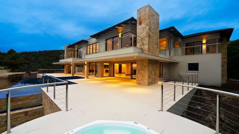 Modern luxury house with large outdoor terrace, pool, and panoramic views, featuring contemporary architecture, stone accents, glass railings, and outdoor lighting at dusk.