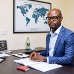 Corporate professional man in formal suit sitting at desk with world map and government emblem in background, representing international business or government leadership.